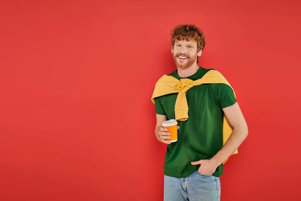 Morning coffee, redhead man with beard and curly hair holding paper cup on coral background, vibrant colors, male fashion, takeaway drink, happy and stylish man posing with hand in pocket — Stock Photo