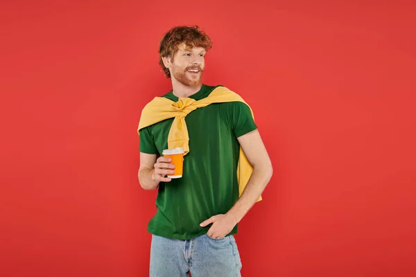 Morning coffee, energy, redhead man with beard and curly hair holding paper cup and standing with hand in pocket, on coral background, vibrant colors, male fashion, takeaway drink, hot beverage — Stock Photo