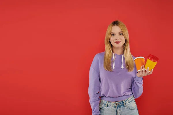 Takeaway drink, youthful fashion, blonde young woman in casual attire holding paper cups on coral background, cheerful, looking at camera, vibrant colors, fashion forward, hot beverage — Stock Photo