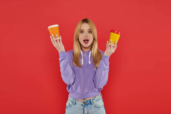 Takeaway drink, youthful fashion, blonde young woman in casual attire holding paper cups on coral background, choice, open mouth, looking at camera, vibrant colors, fashion forward, hot beverage — Stock Photo
