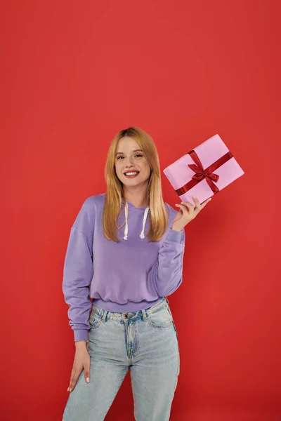 Holiday, positivity, blonde young woman in casual attire holding present on coral background, looking at camera, vibrant colors, wrapped gift box, attractive and stylish, festive occasions — Stock Photo