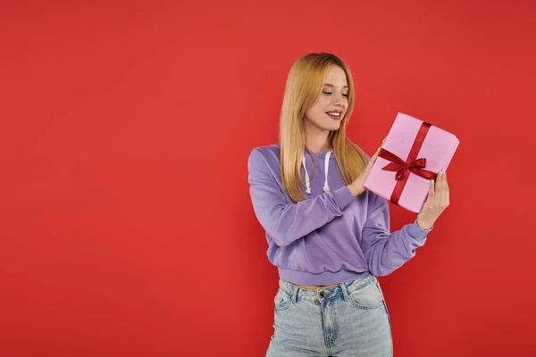 Holiday, cheerfulness, blonde woman in casual attire holding present on coral background, smiling, vibrant colors, wrapped gift box, attractive and stylish, festive occasions — Stock Photo