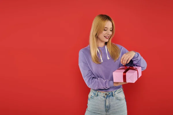 Holiday, happiness, blonde young woman in casual attire opening present on coral background, vibrant colors, wrapped gift box, attractive and stylish, festive occasions, purple hoodie and jeans — Stock Photo