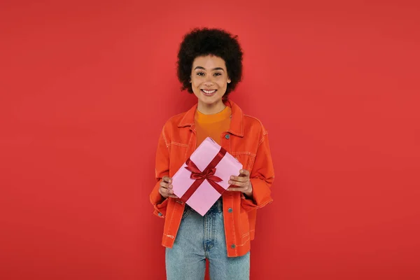 Holiday, happy african american woman in casual attire holding wrapped present on coral background, vibrant colors, gift box, attractive and stylish, festive occasions, looking at camera — Stock Photo