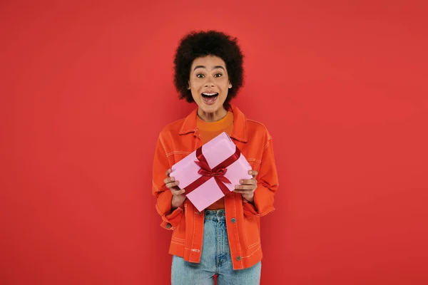 Urlaub, erstaunt afrikanisch-amerikanische Frau in lässiger Kleidung halten verpackt Geschenk auf Korallenhintergrund, lebendige Farben, Geschenkbox, attraktive und stilvolle, festliche Anlässe, Blick in die Kamera — Stockfoto