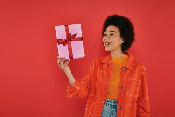 Holiday, excited african american woman in casual attire holding wrapped present on coral background, vibrant colors, gift box, attractive and stylish, festive occasions, trendy look — Stock Photo