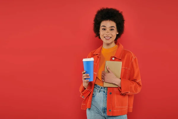 Boisson à emporter, femme afro-américaine gaie en tenue décontractée tenant livre sur fond de corail, couleurs vives, attrayant et élégant, café à emporter, tasse en papier, étudiante regardant la caméra — Photo de stock