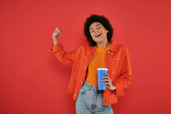 Take-away-Drink, fröhliche afrikanisch-amerikanische Frau in lässiger Kleidung mit Pappbecher auf Korallenhintergrund, lebendige Farben, attraktiv und stilvoll, Coffee to go, aufgeregt und inspiriert — Stockfoto