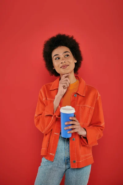 Bebida takeaway, mulher americana africana pensativa e encaracolada em traje casual segurando copo de papel no fundo de coral, cores vibrantes, atraente e elegante, café para ir, copo descartável — Fotografia de Stock