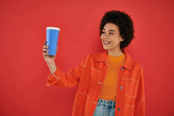 Boisson à emporter, joyeuse femme afro-américaine en tenue décontractée regardant tasse en papier sur fond corail, couleurs vives, attrayant et élégant, café à emporter, tasse jetable avec boisson chaude — Photo de stock