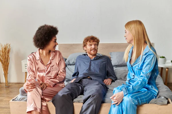 Polyamorous lovers, understanding, three adults, redhead man and multicultural women in pajamas sitting on bed at home, cultural diversity, acceptance, bisexual, positive, open relationship concept — Stock Photo