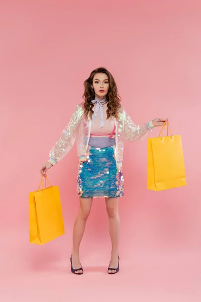 Konsum, schöne Frau posiert mit Einkaufstaschen, steht wie eine Puppe auf rosa Hintergrund, Konzeptfotografie, Puppenpose, stylisches Outfit, Modell im Rock mit Pailletten und transparenter Jacke — Stockfoto