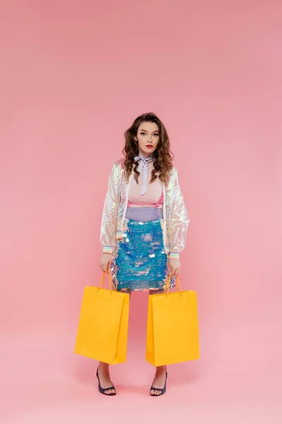 Attraktive Frau mit Einkaufstaschen auf rosa Hintergrund, Konzeptfotografie, Konsumverhalten, stylischem Outfit, junges Model im Rock mit Pailletten und transparenter Jacke blickt in die Kamera — Stockfoto