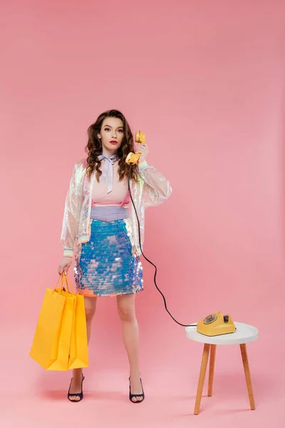 Hermosa mujer joven llevando bolsas de compras, hablando por teléfono retro, de pie como una muñeca sobre fondo rosa, fotografía conceptual, pose muñeca, modelo en falda con lentejuelas y chaqueta transparente - foto de stock