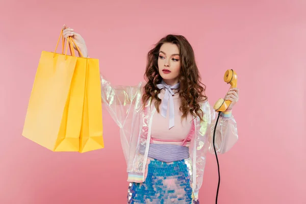 Phone call, attractive woman carrying shopping bags and holding retro handset on pink background, phone, concept photography, consumerism, young model in skirt with sequins and transparent jacket — Stock Photo
