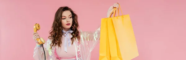 Attractive woman carrying shopping bags and holding retro handset on pink background, vintage phone, concept photography, consumerism, young model in transparent jacket, banner — Stock Photo