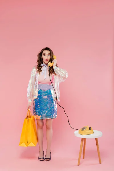 Sorprendida mujer joven llevando bolsas de compras y hablando por teléfono retro, de pie sobre fondo rosa, fotografía conceptual, llamada telefónica, teléfono vintage, concepto de ama de casa - foto de stock