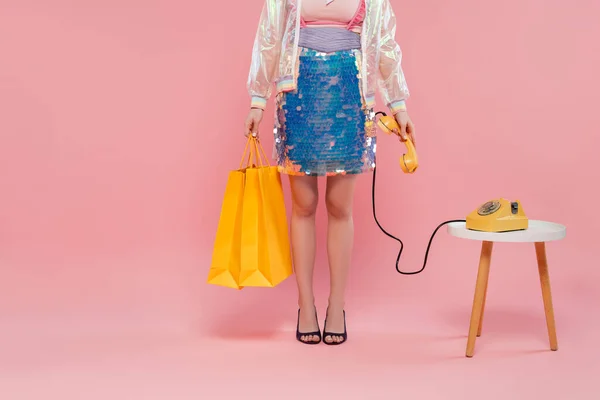 Konzeptfotografie, beschnittene Ansicht einer jungen Frau, die Einkaufstüten trägt und ein Retro-Telefon in der Hand hält, auf rosa Hintergrund stehend, Telefonanruf, Vintage-Telefon, Hausfrauenkonzept — Stockfoto
