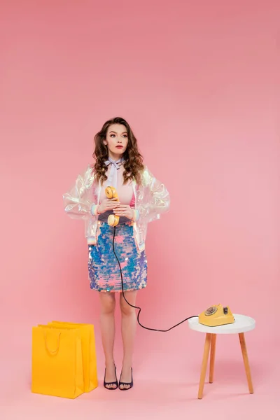 Красивая молодая женщина позирует возле торговых мешков, стоя как кукла и держа ретро телефон на розовом фоне, концептуальная фотография, кукольная поза, стильный наряд, домохозяйка делает телефонный звонок — стоковое фото