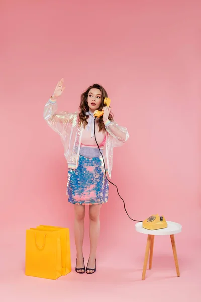 Hermosa mujer que tiene llamada telefónica, posando cerca de bolsas de compras, de pie como una muñeca y sosteniendo el auricular retro sobre fondo rosa, fotografía conceptual, pose de muñeca, ama de casa mirando las uñas - foto de stock
