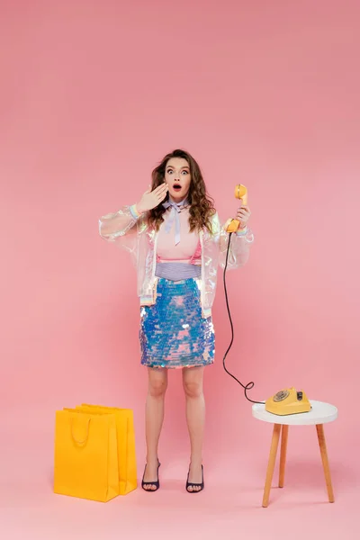 Шокированная женщина позирует возле торговых мешков, стоя как кукла и держа ретро телефон на розовом фоне, концептуальная фотография, кукольная поза, стильный наряд, домохозяйка делает телефонный звонок — стоковое фото