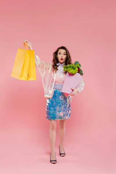 Jovem mulher espantada carregando sacos de compras e saco de supermercado com legumes, de pé sobre fundo rosa, fotografia conceitual, deveres domésticos, conceito dona de casa — Fotografia de Stock