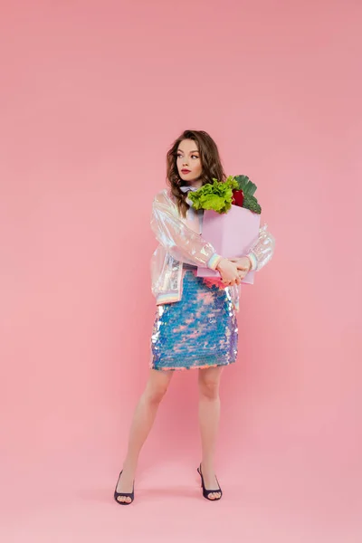 Housewife concept, attractive young woman carrying grocery bag with vegetables, model with wavy hair standing on pink background, conceptual photography, home duties, stylish wife — Stock Photo