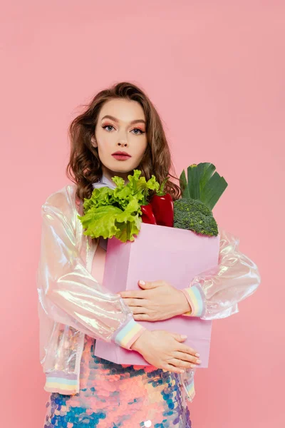 Housewife concept, beautiful young woman carrying grocery bag with vegetables, model with wavy hair standing on pink background, conceptual photography, home duties, stylish wife, portrait — Stock Photo
