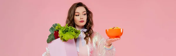 Housewife concept, attractive young woman carrying grocery bag with vegetables and bowl with corn flakes, model with wavy hair on pink background, conceptual photography, home duties, banner — Stock Photo