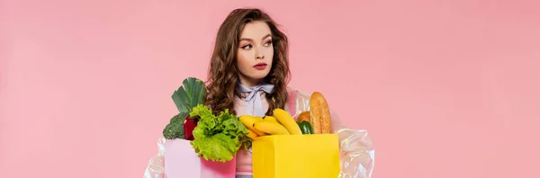 Housewife concept, attractive young woman carrying grocery bags with vegetables and bananas, model with wavy hair on pink background, conceptual photography, home duties, stylish wife, banner — Stock Photo
