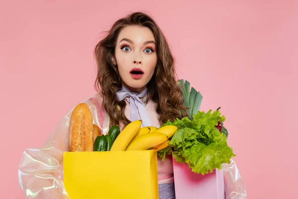 Hausfrauenkonzept, schockierte junge Frau, die Einkaufstüten mit Gemüse und Bananen trägt, Model mit welligem Haar auf rosa Hintergrund, konzeptionelle Fotografie, häusliche Pflichten, stilvolle Ehefrau — Stockfoto