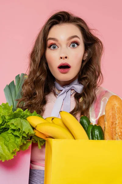 Conceito dona de casa, mulher chocada carregando sacos de supermercado com legumes e bananas, modelo com cabelo ondulado em fundo rosa, fotografia conceitual, deveres domésticos, emocional, retrato — Fotografia de Stock