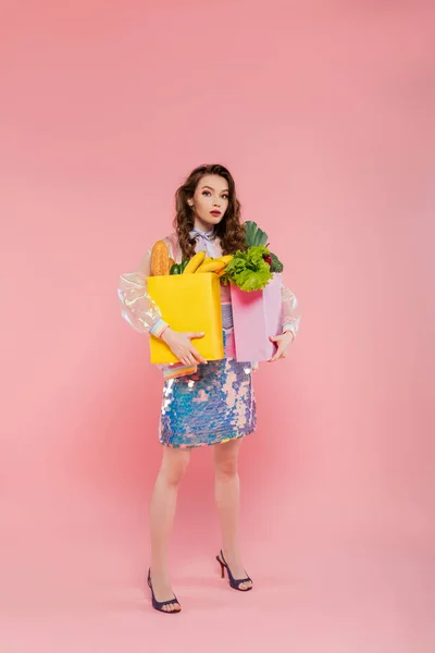 Tâches ménagères, attrayant jeune femme portant des sacs en papier avec épicerie, modèle avec cheveux ondulés debout sur fond rose, photographie conceptuelle, tâches à la maison, femme élégante — Photo de stock