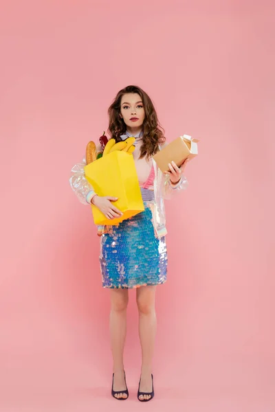 Концепция домохозяйки, привлекательная молодая женщина с сумкой и коробкой с молоком, модель с волнистыми волосами на розовом фоне, концептуальная фотография, домашние обязанности, стильная жена — стоковое фото