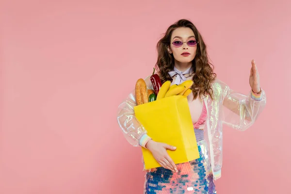 Концепция домохозяйки, молодая женщина в солнечных очках с бумажным пакетом с продуктами, модель брюнетки с волнистыми волосами, позирующая как кукла на розовом фоне, концептуальная фотография, домашние обязанности — стоковое фото