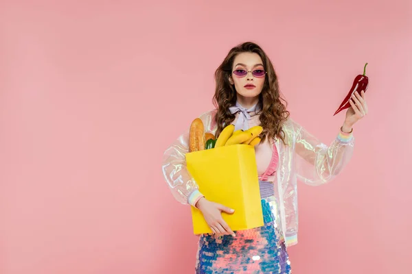 Hausfrauenkonzept, attraktive junge Frau mit Sonnenbrille, Papiertüte mit Lebensmitteln und rotem Pfeffer in der Hand, posiert wie eine Puppe auf rosa Hintergrund, konzeptionelle Fotografie, häusliche Pflichten — Stockfoto
