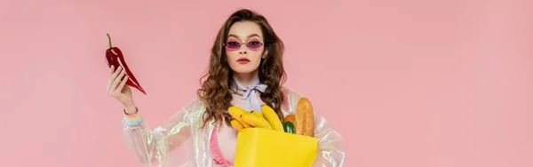 Концепция домохозяйки, привлекательная молодая женщина в солнечных очках с бумажным пакетом с продуктами и с красным перцем в руках, позирует как кукла на розовом фоне, концептуальная фотография, баннер — стоковое фото