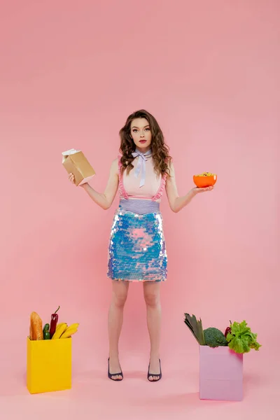 Hausfrauenkonzept, attraktive junge Frau posiert wie eine Puppe, hält Karton mit Milch und Schale mit Cornflakes in der Nähe von Papiertüten mit Lebensmitteln, rosa Hintergrund, konzeptuelle Fotografie — Stockfoto