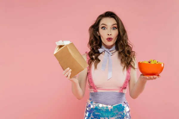 Housewife concept, shocked young woman posing like a doll, holding takeaway food and bowl with corn flakes, pink background, conceptual photography, home duties, emotional — Stock Photo