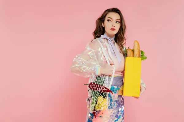 Housewife concept, beautiful young woman holding reusable mesh bag with groceries, stylish wife doing daily house duties, standing on pink background, looking away, role play — Stock Photo