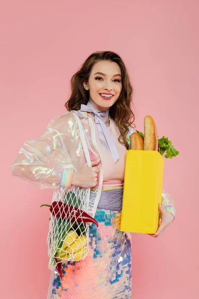 Housewife concept, happy young woman holding reusable mesh bag with groceries, stylish wife doing daily house duties, standing on pink background, looking at camera, role play — Stock Photo