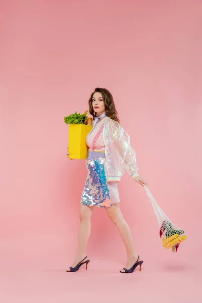 Концепция домохозяйки, счастливая молодая женщина с многоразовой веревочной сумкой с продуктовыми наборами, стильная жена, выполняющая ежедневные домашние обязанности, ходящая на розовом фоне, смотрящая на камеру, модная — стоковое фото