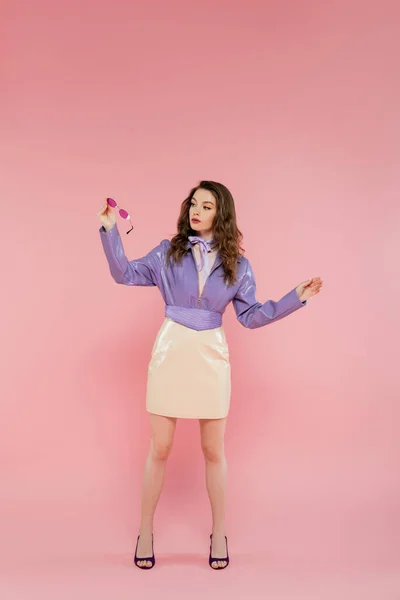 Glamour, beautiful young woman gesturing and looking at sunglasses, acting like a doll, fashionable outfit, model in purple jacket and skirt, pink background, studio shot, conceptual, full length — Stock Photo