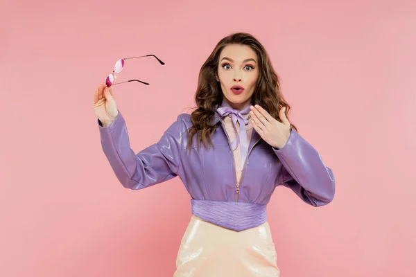 Glamour, surprised young woman gesturing and looking at camera, holding sunglasses, fashionable outfit, model in purple jacket and skirt standing on pink background, studio shot, acting like a doll — Stock Photo