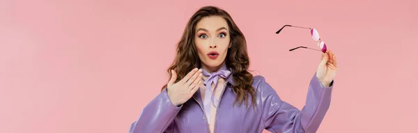 Glamour, surprised young woman gesturing and looking at camera, holding sunglasses, trendy outfit, model in purple jacket standing on pink background, studio shot, conceptual, banner — Stock Photo