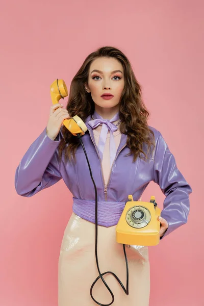 Концепція ляльки, молода жінка з хвилястим волоссям тримає телефон і ретро, модний одяг, брюнетка модель в фіолетовій куртці позує і дивиться на камеру на рожевому фоні, студійний знімок — стокове фото