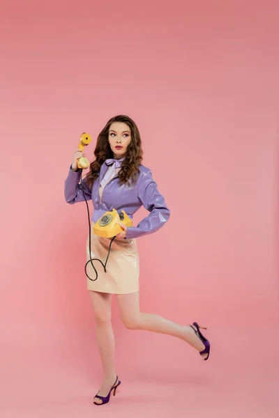 Concepto de muñeca, mujer joven con el pelo ondulado sosteniendo el teléfono y el teléfono retro amarillo, traje de moda, modelo morena en chaqueta púrpura posando y mirando hacia otro lado sobre fondo rosa, plano de estudio, longitud completa - foto de stock