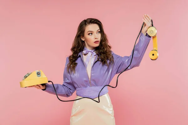 Pose muñeca, hermosa mujer joven con el pelo ondulado mirando el teléfono mientras sostiene el teléfono retro amarillo, modelo morena en chaqueta púrpura posando sobre fondo rosa, tiro al estudio - foto de stock