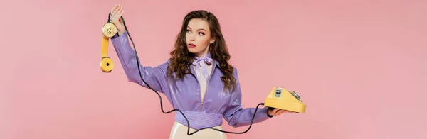 Pose muñeca, hermosa mujer joven con el pelo ondulado mirando el teléfono mientras sostiene el teléfono retro amarillo, modelo morena en chaqueta púrpura posando sobre fondo rosa, tiro al estudio, pancarta - foto de stock