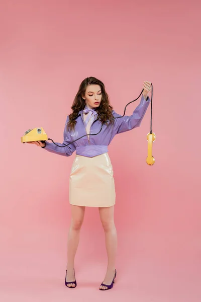 Puppen-Pose, schöne junge Frau mit welligem Haar, die auf Handy schaut, während sie gelbes Retro-Telefon hält, brünettes Modell in lila Lederjacke, das auf rosa Hintergrund posiert, Studioaufnahme, volle Länge — Stockfoto